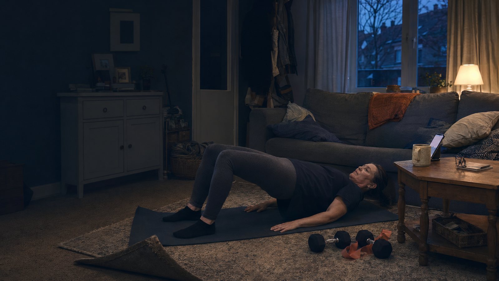 A woman training on a mat in her front room at dusk, with dumbbells and a resistance band beside her.